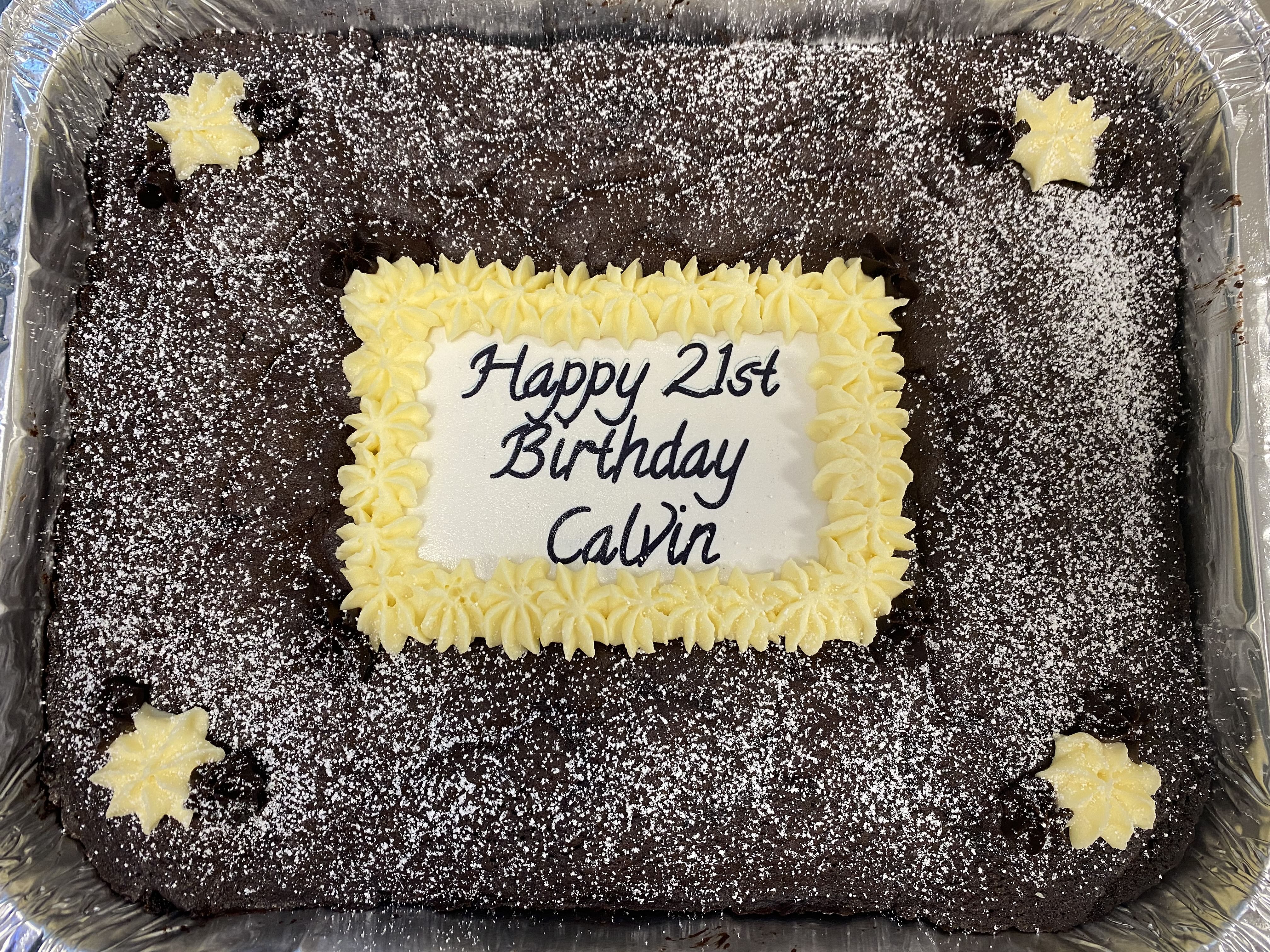 A rectangular silver tray of uncut chocolate brownies is decorated with powdered sugar, small yellow star shapes in the corners, and a white icing rectangle in the center. The icing reads 'Happy 21st Birthday Calvin' in black letters, capturing an old-fashioned recipe's charm with its rich chocolatey flavor.